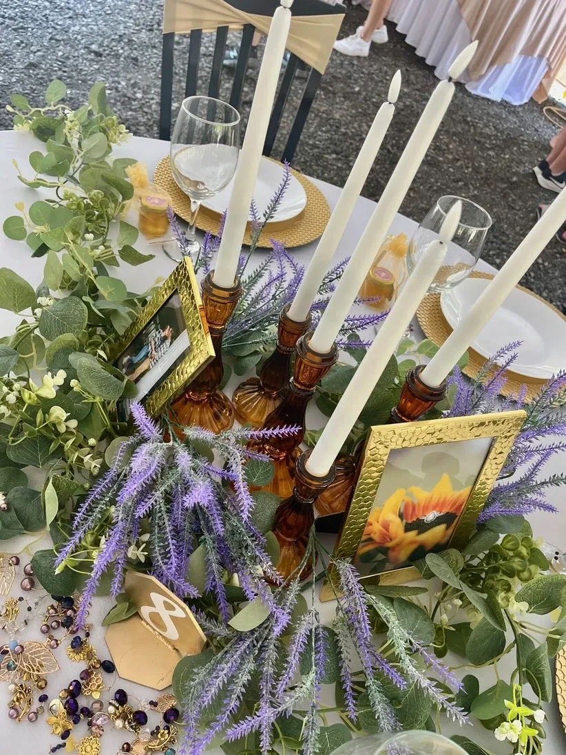 download (1) Wedding table centerpiece at Riverbend Events in Weedsport, NY, featuring amber glass candlesticks with white taper candles, gold photo frames, lush greenery and purple floral accents, elegant place settings with gold chargers, wine glasses, and a wooden table number eight.