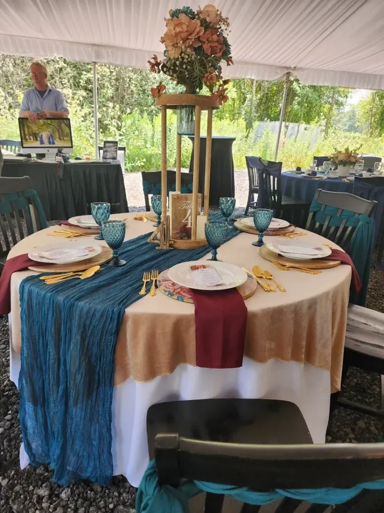 Elegantly decorated round wedding reception table at Riverbend Events in Weedsport, NY, featuring layered beige and white tablecloths with a blue runner, gold flatware, blue goblets, burgundy napkins, floral centerpiece on a wooden stand, surrounded by black chairs with blue sashes under a tent showcasing natural outdoor views.