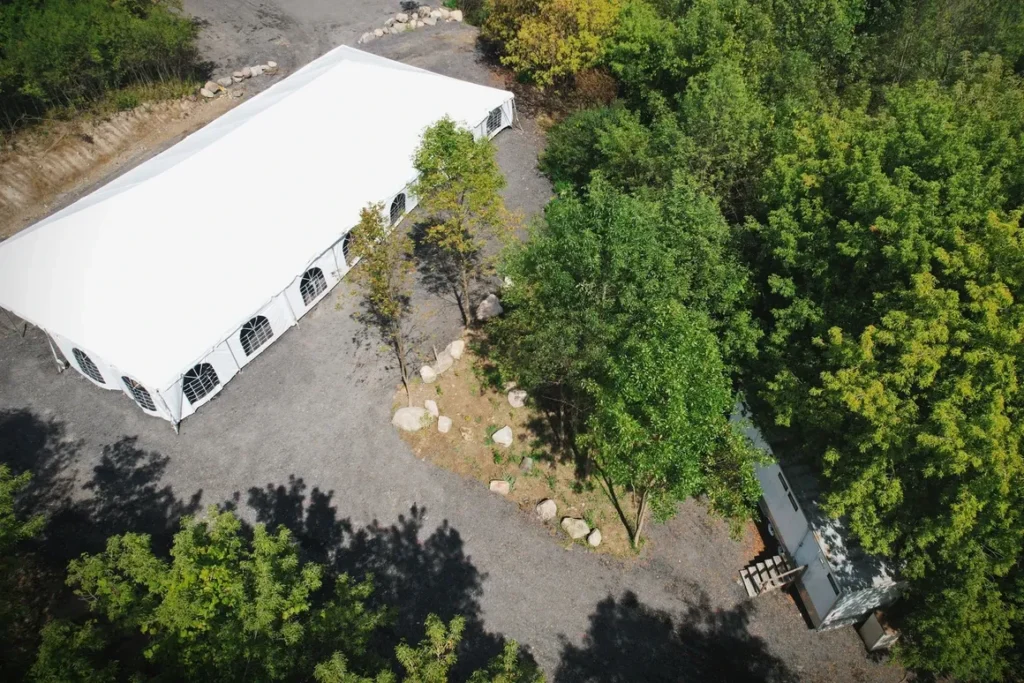Aerial view of a large outdoor wedding tent set on gravel surrounded by lush green trees and natural landscape, showcasing a serene and elegant venue space for weddings and special occasions.