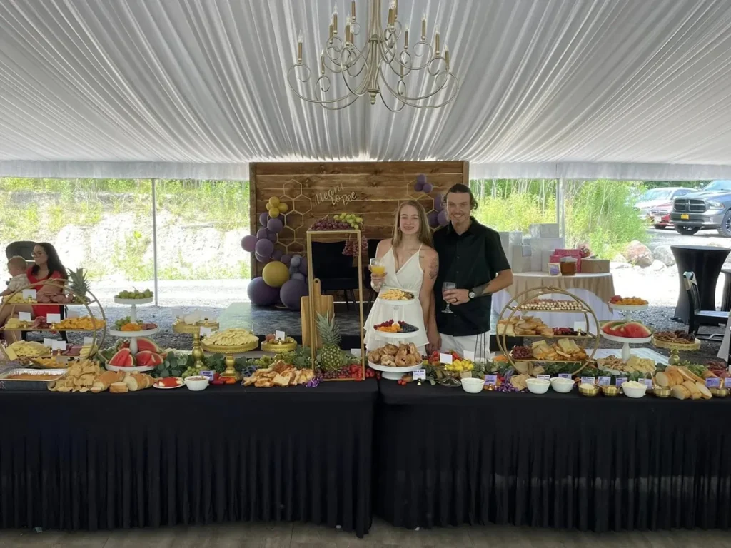 Couple standing behind an elegant buffet table with assorted cheeses, fruits, pastries, and appetizers under a white draped tent showcasing a personalized and natural setting for special occasions.