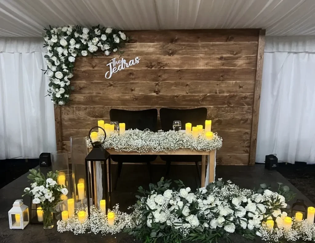 Rustic wooden wedding backdrop with white floral arrangements, baby's breath, glowing candles, and a personalized sign reading 