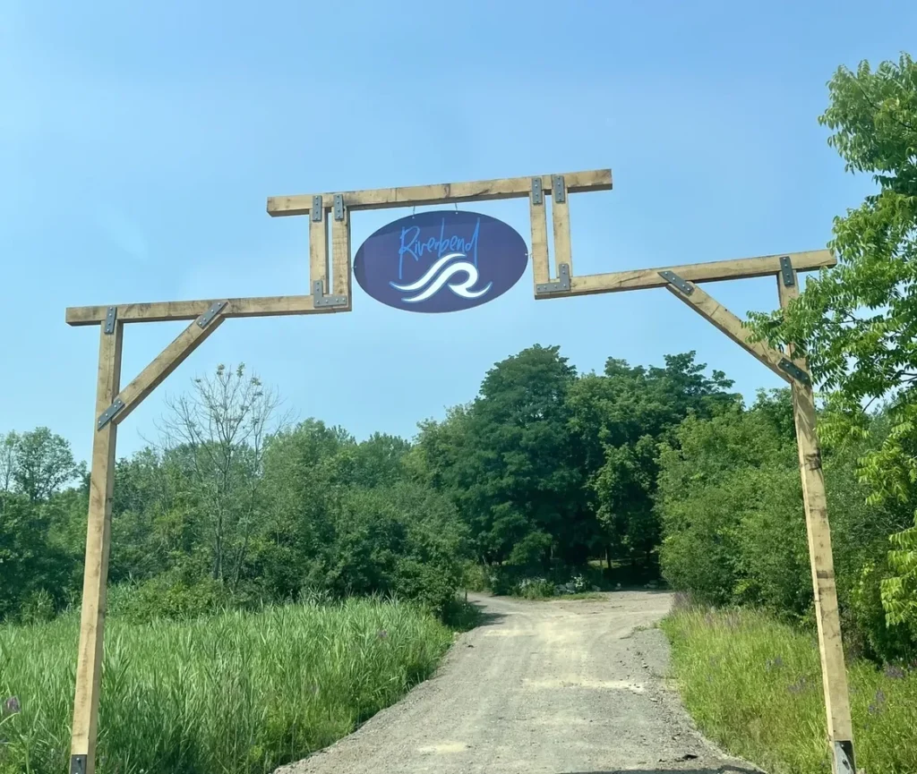 Entrance archway with a wooden frame and a hanging Riverbend Events sign above a gravel driveway surrounded by lush green trees and grass, showcasing the natural beauty of the Weedsport, NY wedding venue.