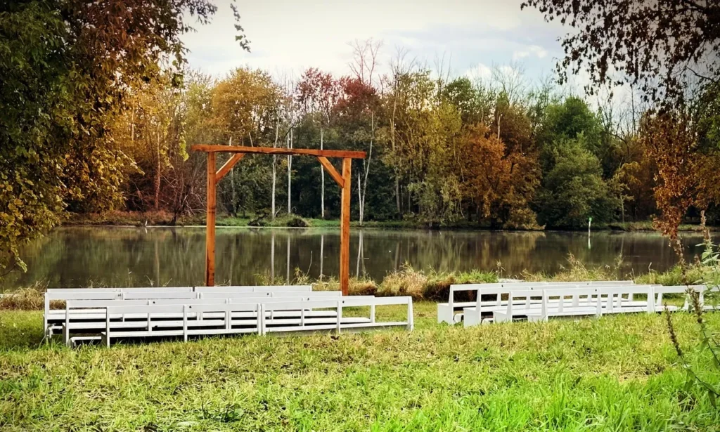 Outdoor wedding ceremony setup with white benches and a wooden arch by a calm river, surrounded by trees with autumn foliage at Riverbend Events in Weedsport, NY