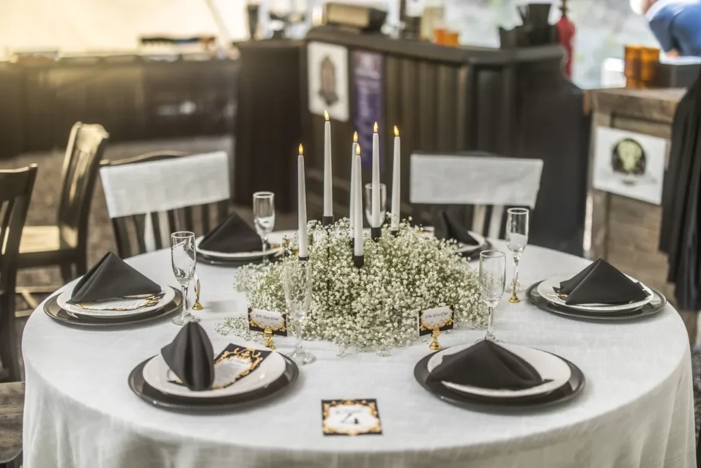 Round wedding reception table set with white tablecloth, black napkins, champagne flutes, a centerpiece of white flowers and tall candles, showcasing elegant and intimate event decor