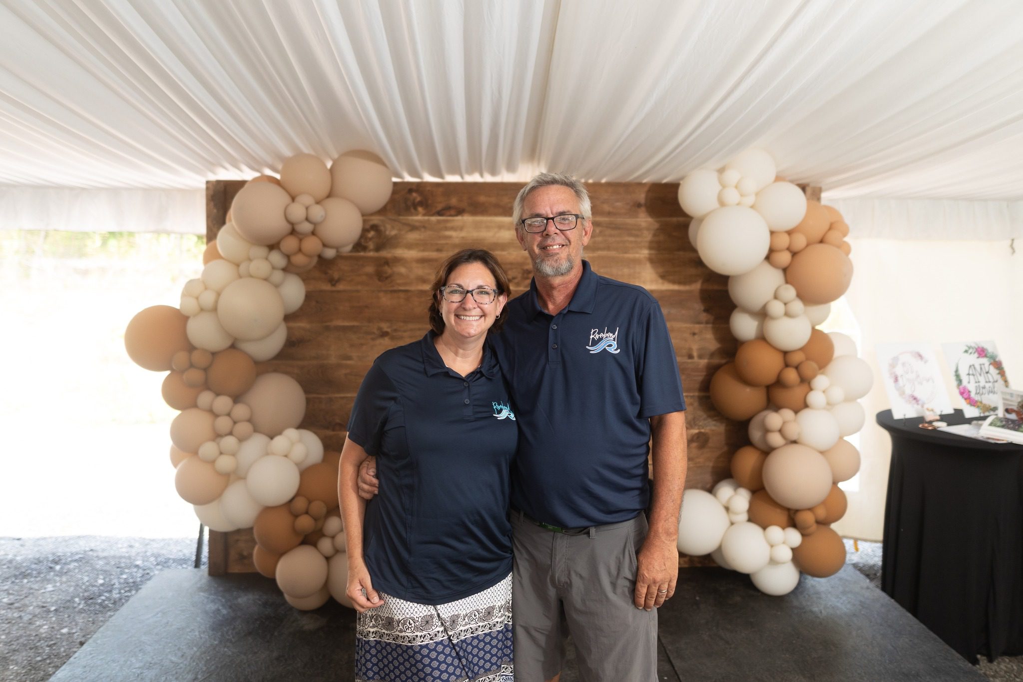 team Mandy and Greg standing together smiling under a white tent with beige and brown balloon arches on either side and a wooden backdrop, wearing matching navy Riverbend Events polo shirts, representing love-filled, personalized wedding and event venue founders.