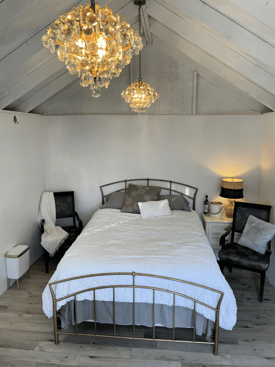 thumbnail_IMG_6246 Cozy bridal cabin bedroom with a metal bed frame, white bedding, decorative pillows, two black chairs with cushions, bedside table with lamp and wine set, under rustic vaulted ceiling with crystal chandeliers at Riverbend Events wedding venue.